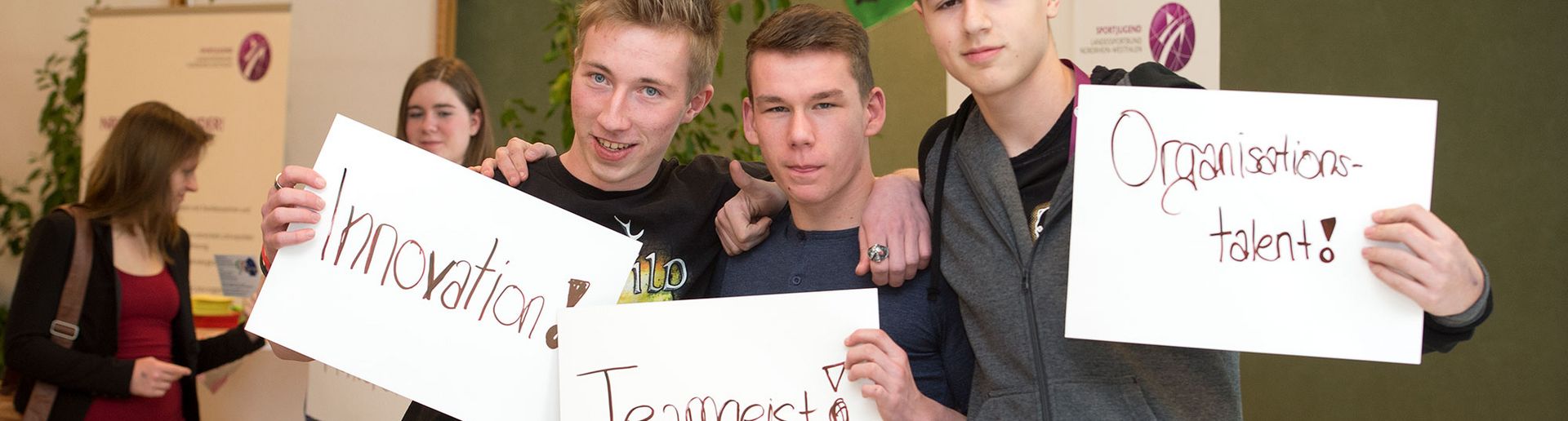 drei Jungs die jewals ein Schild hochhalten . Auf dem einen steht "Innovation", auf dem anderen "Teamsgeist" und auf dem dritten "Organisationstalent"